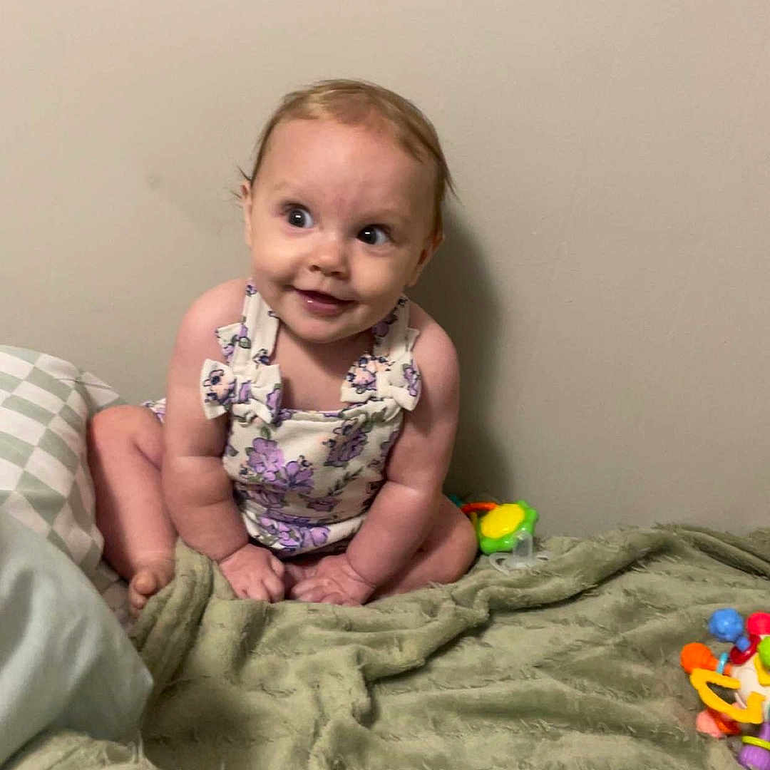 Sophia joined the competition — help win amazing prizes! baby, blanket, child, clothing, colorful, cute, face, flower_pattern, happy, head, indoor, infant, person, playful, sitting, skin, smiling, toy, wall, young