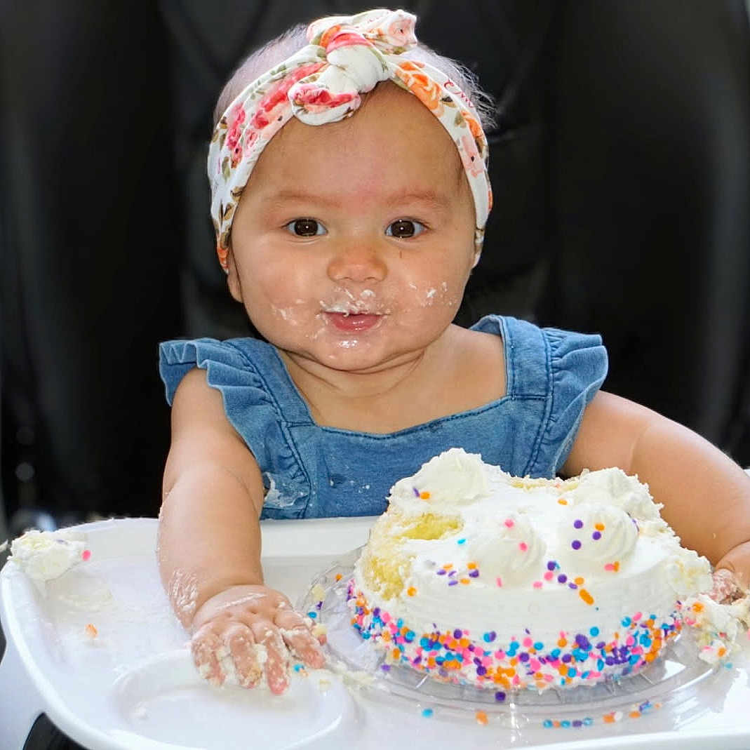 Emmy joined the competition — help win amazing prizes! baby, child, cake, high_chair, sprinkles, frosting, blue_dress, headband, floral, smiling, messy, celebration, indoor, food, birthday, happy, face, hands, person, cute
