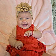 Emmy is registered to the contest to win money with this photo: baby, smiling, headband, orange_dress, blanket, cute, infant, happy, child, lying_down, soft_texture, indoors, adorable, portrait, person, skin, hand, face, head, baby_girl