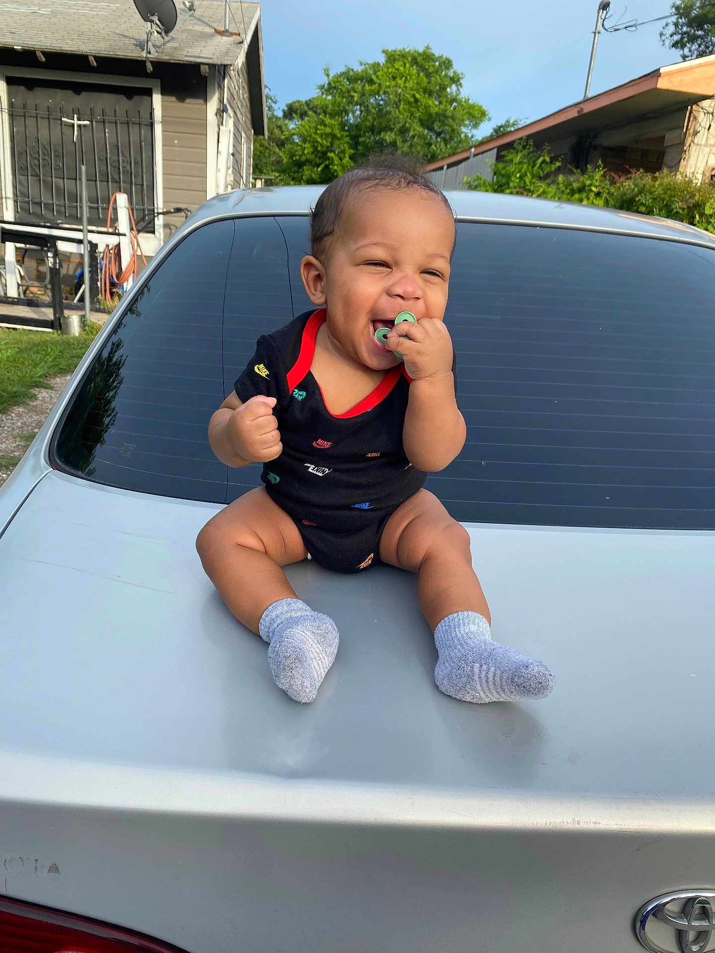 Joseph is registered to the contest to win money with this photo: baby, smiling, pacifier, black_onesie, socks, car_trunk, silver_car, outdoor, daylight, house, greenery, happy, infant, cute, person, casual_clothing, sitting, child, young, front_view