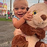 Wyatt is registered to the contest to win money with this photo: toddler, child, plush_toy, stuffed_animal, smiling, happy, outdoor, carnival, fair, toy, person, holding, face, head, clothing, grass, road, pole, sky, festival