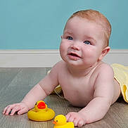 Luca joined the competition — help win amazing prizes! baby, child, rubber_duck, yellow_duck, floor, wooden_floor, towel, blue_wall, skin, face, hands, smile, indoor, cute, infant, play, toy, lying_down, childhood, happy