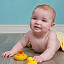 baby, child, rubber_duck, yellow_duck, floor, wooden_floor, towel, blue_wall, skin, face, hands, smile, indoor, cute, infant, play, toy, lying_down, childhood, happy