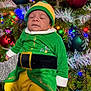 baby, child, sleeping, christmas_tree, ornaments, holiday, festive, elf_costume, green_clothing, yellow_pants, red_ornaments, lights, pine_tree, seasonal, celebration, festivity, cute, holiday_decor, infant, costume