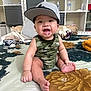 baby, bedspread, camo_clothing, cap, child, drooling, fabric, floral_pattern, foot, hand, happy, indoor, person, room, shelf, sitting, smiling, sneaker, toy, water_bottle