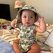 Drake is registered to the contest to win money with this photo: baby, infant, child, hat, sitting, bed, bedding, lamp, indoor, cute, clothing, portrait, face, feet, person, animal_print, curious, soft_lighting, home, room