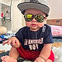 baby, child, sunglasses, cap, drool, sitting, indoor, bedspread, clothing, red_shorts, tshirt, reflection, face, foot, hand, person, cute, casual, expression, home