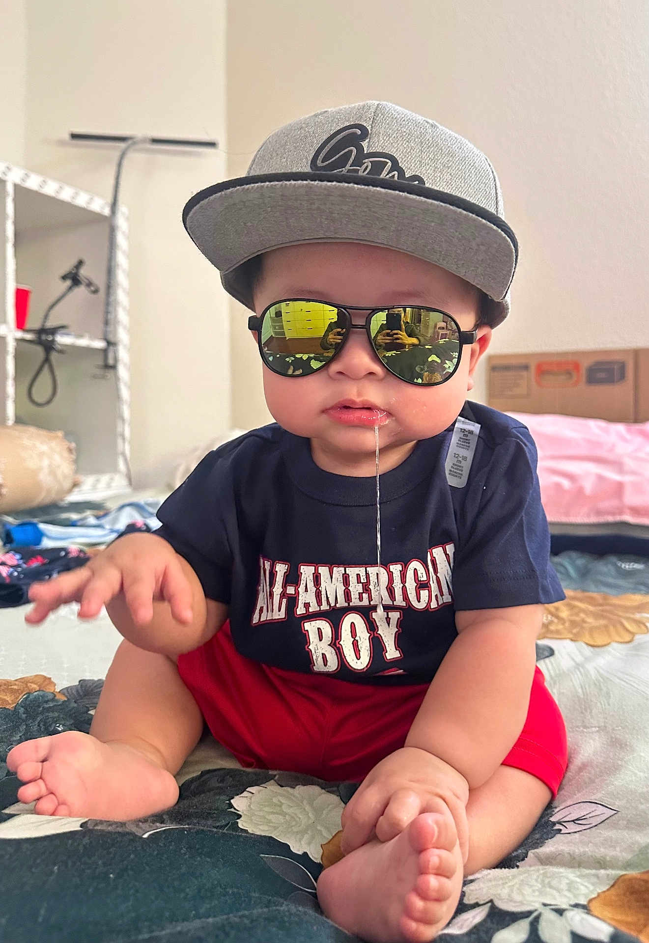 Drake joined the competition — help win amazing prizes! baby, child, sunglasses, cap, drool, sitting, indoor, bedspread, clothing, red_shorts, tshirt, reflection, face, foot, hand, person, cute, casual, expression, home
