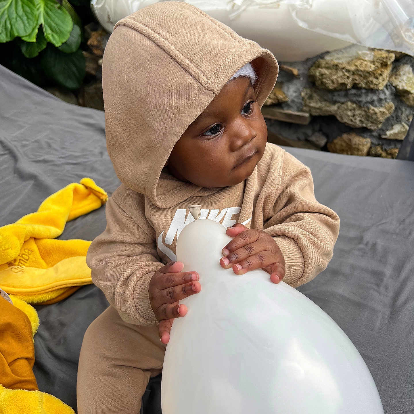 Mamady a rejoint le concours — aidez-le/la à gagner de superbes lots ! toddler, child, hoodie, balloon, yellow_plush_toy, outdoor, greenery, stone, water_feature, gray_surface, curious, clothing, footwear, sitting, portrait, baby, cute, casual, daylight, playful
