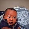 baby, infant, child, portrait, face, eyes, wide_eyed, indoor, bedroom, blanket, bedding, fish_pattern, clothing, dark_shirt, sitting, looking_at_camera, television, home, closeup, photography
