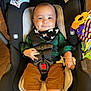 baby, child, car_seat, straps, pacifier, toy, plaid_shirt, bib, dimples, smile, indoor, wooden_floor, comfort, infant, cute, black_straps, green_clothing, brown_pants, colorful_toy, sitting