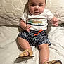 baby, child, infant, sitting, bed, pillow, white_tshirt, shorts, sandals, cute, chubby_cheeks, indoors, smiling, portrait, clothing, footwear, person, young_child, happy, casual