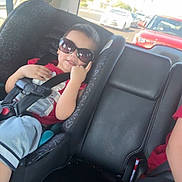 Cylus is registered to the contest to win money with this photo: child, toddler, car_seat, sunglasses, car_interior, seatbelt, smiling, red_shirt, shorts, road, car, traffic, window, daylight, seat, leather, safety, cute, baby, travel