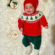 Nicolas is registered to the contest to win money with this photo: baby, child, smiling, red_hat, red_sweater, christmas_sweater, green_pants, white_socks, holiday, christmas, ornament, wooden_decoration, fluffy_rug, indoor, festive, cute, portrait, person, smile, holiday_decor