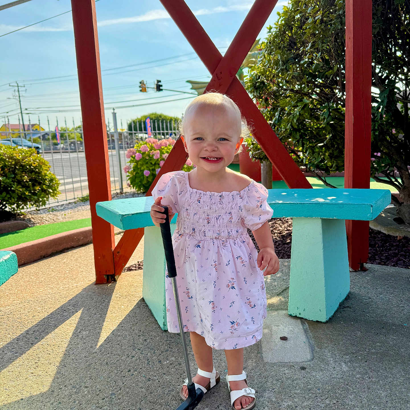 Blaklee joined the competition — help win amazing prizes! bench, bushes, child, concrete, daytime, dress, floral_pattern, flowers, girl, greenery, happy, mini_golf, outdoor, person, playground, putter, sandals, smiling, sunlight, toddler