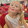 Presley is registered to the contest to win money with this photo: child, girl, blonde_hair, curly_hair, white_dress, red_chair, gymnasium, indoor, seated, playful_expression, number_tag, event, people_in_background, smile, young_child, person, casual, fun, curious, audience