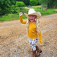 Presley is registered to the contest to win money with this photo: child, cowgirl, cowboy_hat, pigtails, long_hair, smiling, yellow_shirt, leggings, horse_print, boots, gravel_road, outdoor, trees, fence, rural, nature, happy, person, standing, daytime