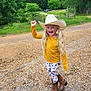 child, cowgirl, cowboy_hat, pigtails, long_hair, smiling, yellow_shirt, leggings, horse_print, boots, gravel_road, outdoor, trees, fence, rural, nature, happy, person, standing, daytime