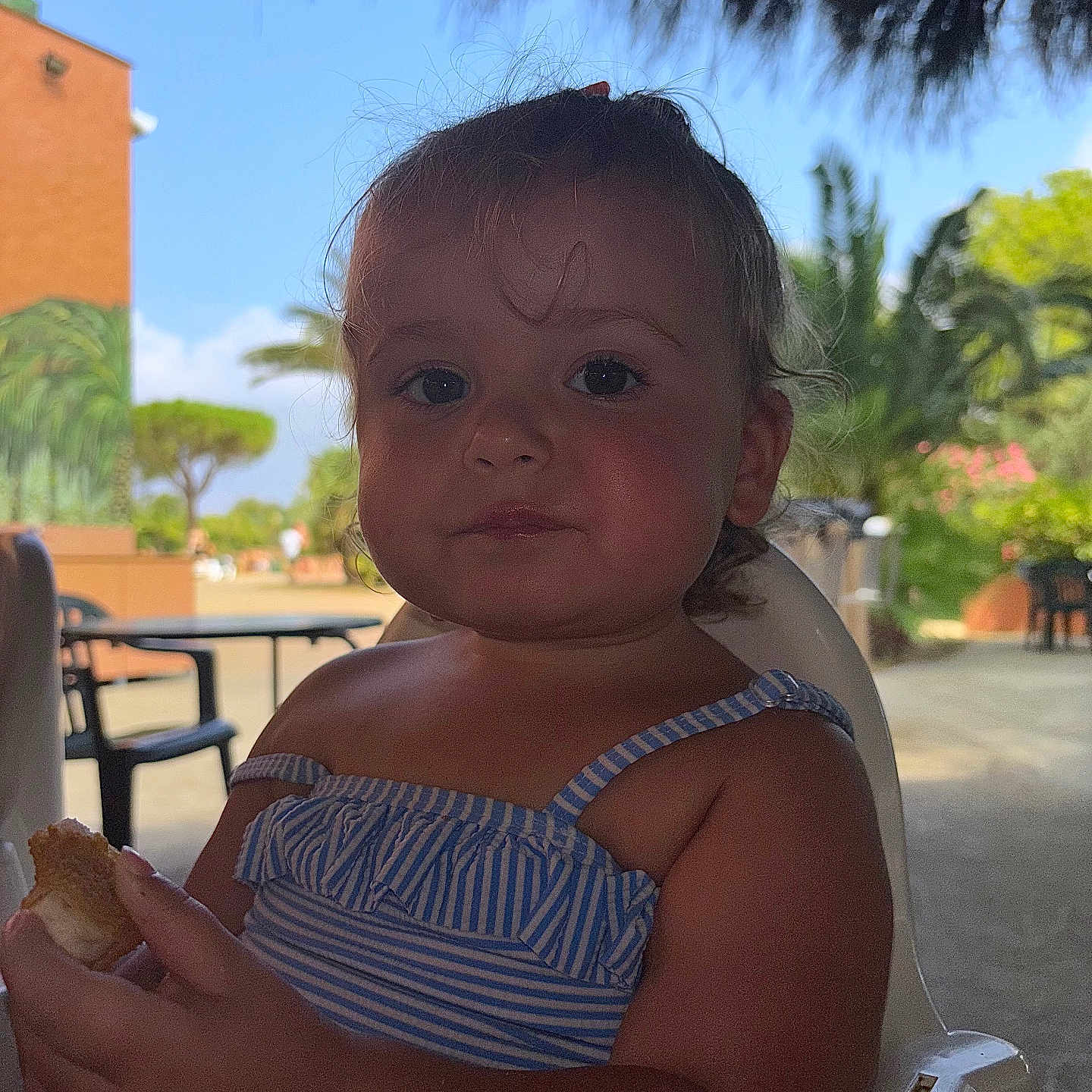 Inaya Delfolie a rejoint le concours — aidez-le/la à gagner de superbes lots ! baby, blue_striped_clothing, bread, child, cute, daylight, eating, food, greenery, nature, outdoor, palm_trees, person, plastic_chair, portrait, relaxed, summer, thatched_roof, toddler, vacation