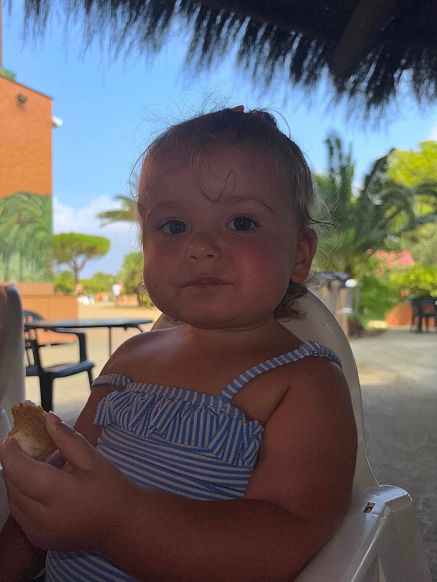 Inaya Delfolie a rejoint le concours — aidez-le/la à gagner de superbes lots ! child, toddler, bread, food, blue_striped_clothing, plastic_chair, outdoor, daylight, palm_trees, thatched_roof, summer, vacation, cute, portrait, baby, person, nature, greenery, relaxed, eating