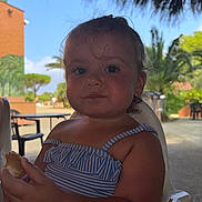 Inaya participe au concours pour gagner de l'argent avec cette photo : child, toddler, bread, food, blue_striped_clothing, plastic_chair, outdoor, daylight, palm_trees, thatched_roof, summer, vacation, cute, portrait, baby, person, nature, greenery, relaxed, eating