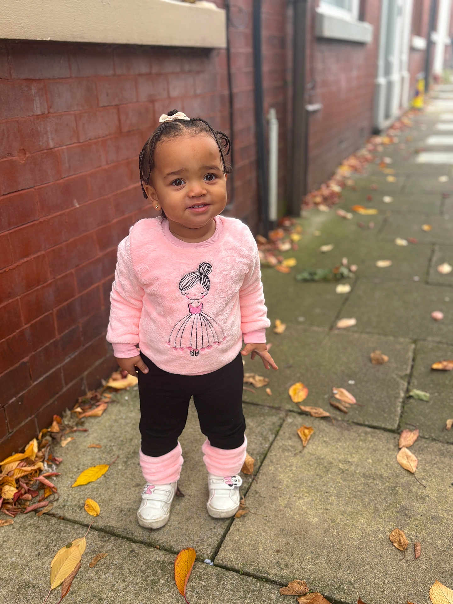Olivia is registered to the contest to win money with this photo: child, toddler, outdoor, sidewalk, brick_wall, fall_leaves, pink_sweater, black_pants, white_shoes, braided_hair, bow, curious_expression, standing, casual_clothing, urban, daylight, person, cute, smile, young_child