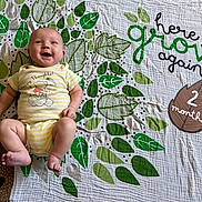 Kaiser joined the competition — help win amazing prizes! baby, blanket, brown, child, cute, green, happy, indoors, infant, leaf_pattern, lying_down, milestone, patterned_fabric, playful, portrait, smiling, text_overlay, two_months, white, yellow_onesie