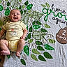 baby, blanket, brown, child, cute, green, happy, indoors, infant, leaf_pattern, lying_down, milestone, patterned_fabric, playful, portrait, smiling, text_overlay, two_months, white, yellow_onesie