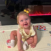 Zoey joined the competition — help win amazing prizes! baby, child, smiling, sitting, yellow_bow, play_mat, electric_fireplace, controller, reflection, indoor, toy, socks, bracelet, face, person, floor, happy, light, hair, clothing