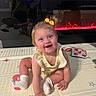 baby, child, smiling, sitting, yellow_bow, play_mat, electric_fireplace, controller, reflection, indoor, toy, socks, bracelet, face, person, floor, happy, light, hair, clothing