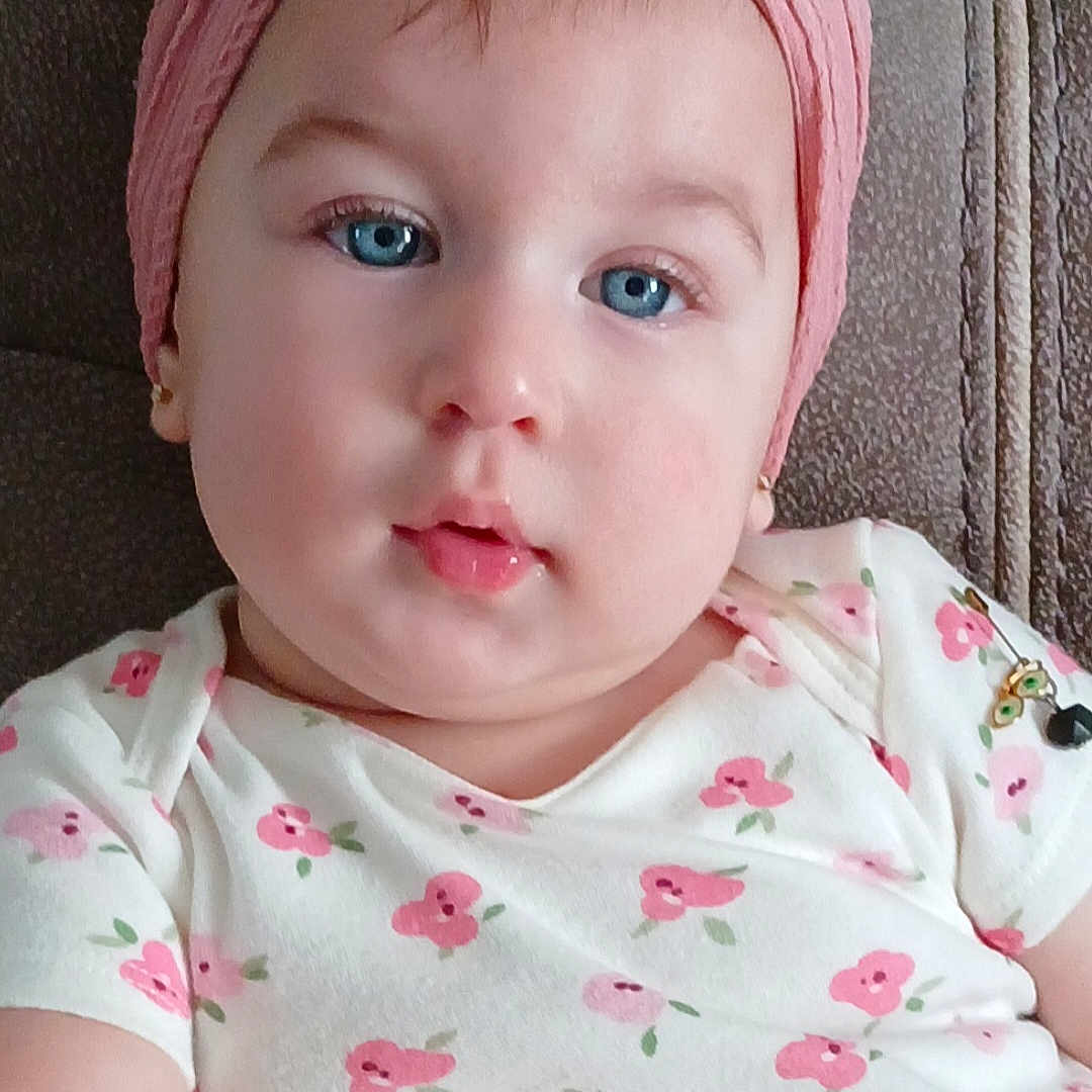 Venus is registered to the contest to win money with this photo: baby, child, face, blue_eyes, headband, flower, pink, white_clothing, floral_pattern, cute, portrait, chubby_cheeks, sitting, indoors, soft_light, skin, earrings, young_child, infant, comfortable