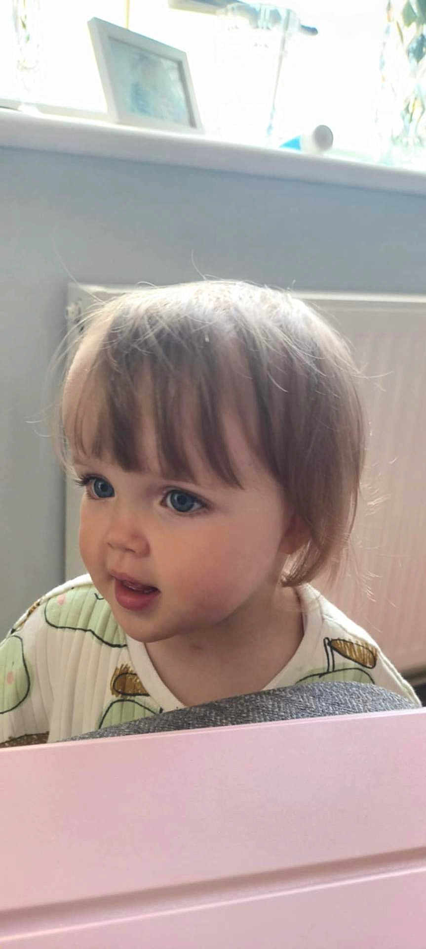 Amelia is registered to the contest to win money with this photo: baby, blue_eyes, child, clothing, curious, cute, expression, face, framed_photo, hair, indoors, looking, patterned_outfit, person, pink_surface, portrait, radiator, soft_lighting, toddler, young_child