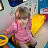 amusement, backpack, blue_eyes, blue_ride, child, clothing, colorful, curious_expression, footwear, ice_cream_drawings, indoor, kiddie_ride, person, pink_jacket, plastic, playful, portrait, toddler, toy, yellow_seat