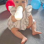 Amelia is registered to the contest to win money with this photo: baby, child, bottle, milk, blue_eyes, dress, carpet, balloons, indoor, sitting, cute, person, infant, milk_bottle, floor, play_area, happy, light, home, young_child