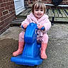 blue_toy, brick_wall, casual, child, concrete, cute, daylight, fun, happy, outdoor, person, pink_coat, play, rain_boots, rocking_horse, smile, toddler, toy, wooden_deck, young_child