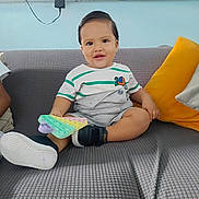 Noah is registered to the contest to win money with this photo: baby, child, toddler, smile, sitting, sofa, couch, cushion, pillow, toy, pop_it_toy, sneakers, shoe, striped_shirt, onesie, happy, portrait, living_room, wall_socket, charger