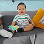 baby, child, toddler, smile, sitting, sofa, couch, cushion, pillow, toy, pop_it_toy, sneakers, shoe, striped_shirt, onesie, happy, portrait, living_room, wall_socket, charger