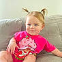 blue_eyes, cartoon_print, casual, child, couch, cute, happy, home, indoor, light, pigtails, pink_shirt, portrait, relaxed, seated, short_sleeves, skin, smiling, toddler, young_child