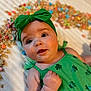 Paisley joined the competition — help win amazing prizes! baby, child, green_headband, green_dress, sparkly_clothing, cereal, colorful, white_blanket, cute, happy, portrait, infant, cheerful, indoors, closeup, smiling, playful, food, background_blur, hand