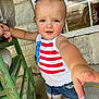 Brooklynn is registered to the contest to win money with this photo: toddler, child, barefoot, outdoor, summer_clothing, denim_shorts, tank_top, american_flag, playful, reaching_hand, green_chair, concrete_floor, stone_wall, window, reflection, hair_bow, person, casual, daylight, portrait