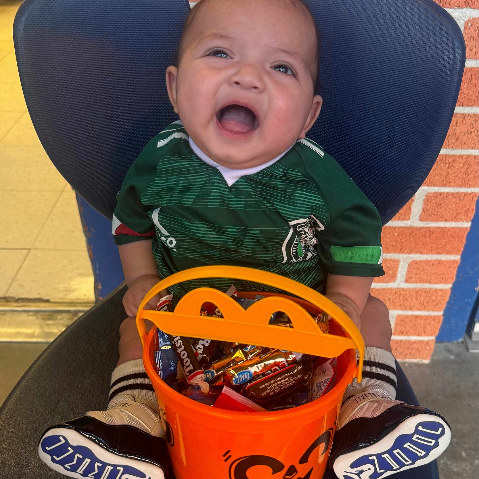 Miguelangel joined the competition — help win amazing prizes! baby, brick_wall, candy, celebration, chair, child, cute, festive, green_jersey, halloween, happy, indoor, joy, orange_bucket, person, portrait, smiling, sneakers, soccer_shirt, socks