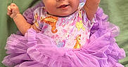 Sienna is registered to the contest to win money with this photo: baby, infant, child, portrait, smiling, happy, bow, headband, tutu, dress, purple, floral, ruffles, fist, raised_arm, green_blanket, lying_down, indoor, colorful_clothing, celebration