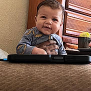 George is registered to the contest to win money with this photo: baby, infant, child, smiling, face, hand, adult_hand, onesie, patterned_clothing, wooden_dresser, mug, nightstand, phone, electronic_device, cozy, bedroom, wall_texture, portrait, sitting, cute