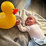baby, baby_toy, blanket, child, comfort, curious, cute, floor, home, indoors, infant, laying_down, playful, rubber_duck, small_child, soft_texture, toy, warm_lighting, white_clothing, wooden_floor