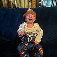 Geremiah is registered to the contest to win money with this photo: baby, barefoot, blue_couch, casual, child, clothing, couch, face, football_pattern, happy, hat, home, indoor, infant, long_sleeve_shirt, pants, person, sitting, smile, wooden_stairs