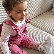 Kayna a rejoint le concours — aidez-le/la à gagner de superbes lots ! toddler, child, curly_hair, pink_overalls, white_shoes, sitting, couch, indoor, cute, happy, fashion, portrait, young_child, smile, comfortable, casual_clothing, home, cozy, striped_couch, curious