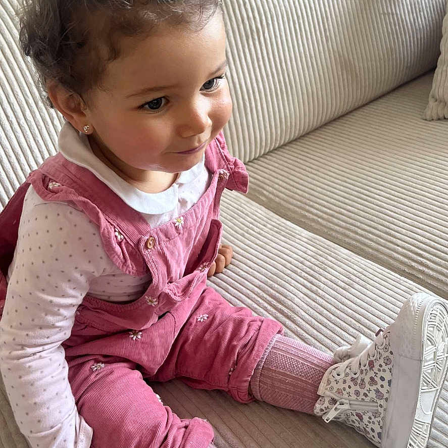 Kayna a rejoint le concours — aidez-le/la à gagner de superbes lots ! toddler, child, curly_hair, pink_overalls, white_shoes, sitting, couch, indoor, cute, happy, fashion, portrait, young_child, smile, comfortable, casual_clothing, home, cozy, striped_couch, curious