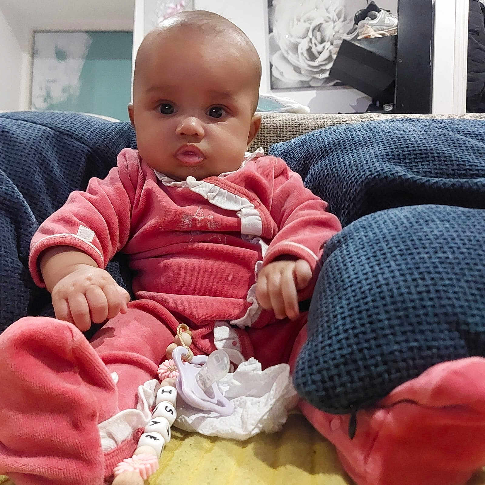 Isaé participe au concours pour gagner de l'argent avec cette photo : baby, beads, blanket, blue_cushion, chair, child, cute, decor, expression, face, footwear, hand, home, indoor, infant, pacifier, pink_clothing, portrait, shelf, sitting