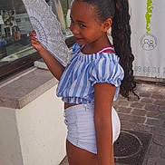 Tyana Wautier participe au concours pour gagner de l'argent avec cette photo : child, girl, fan, blue_striped_top, white_shorts, white_sneakers, long_hair, curly_hair, outdoor, side_view, smiling, street, cobblestone, window, reflection, building, casual_clothing, daylight, fashion, portrait
