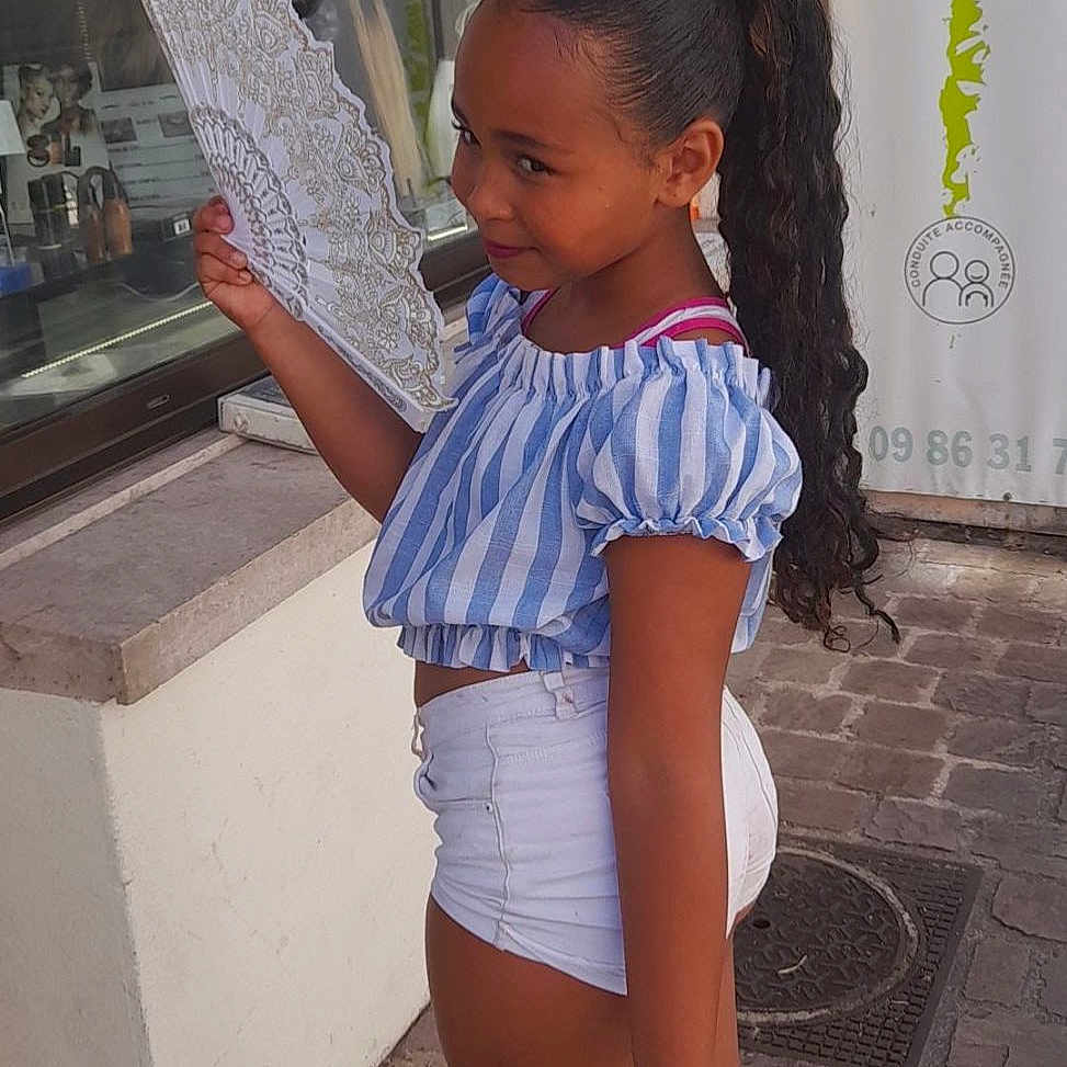 Tyana Wautier participe au concours pour gagner de l'argent avec cette photo : blue_striped_top, building, casual_clothing, child, cobblestone, curly_hair, daylight, fan, fashion, girl, long_hair, outdoor, portrait, reflection, side_view, smiling, street, white_shorts, white_sneakers, window